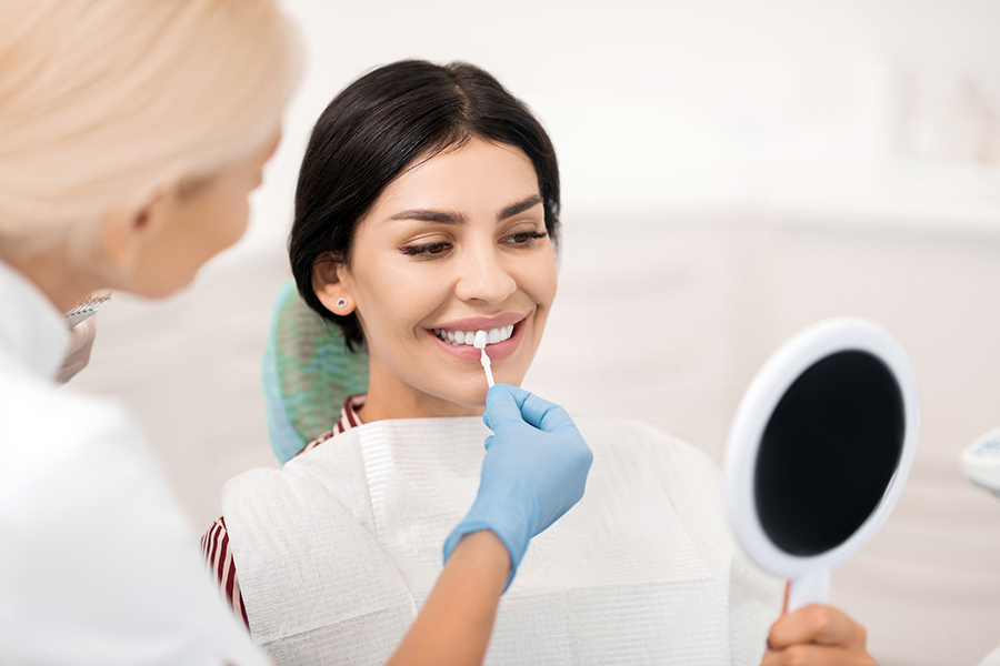 Woman trying on teeth whitening sample at dentists | Butler Family Dental Dental clinician in gloves applies a treatment to a smiling patient’s lips with a cotton swab, patient holding a mirror nearby.